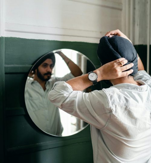 Confident bearded Sikh male wearing denim shirt adjusting traditional headwear standing in front of mirror on wall at home while taking care of appearance